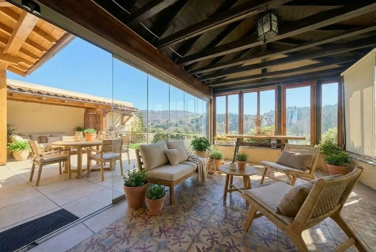 A sunroom with glass walls and a dark wood ceiling features wicker furniture, potted plants, and patterned tile flooring.