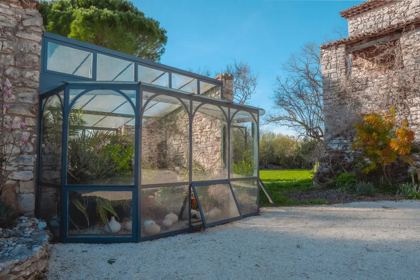 A greenhouse with a dark frame sits on a gravel path next to a stone building under a blue sky.