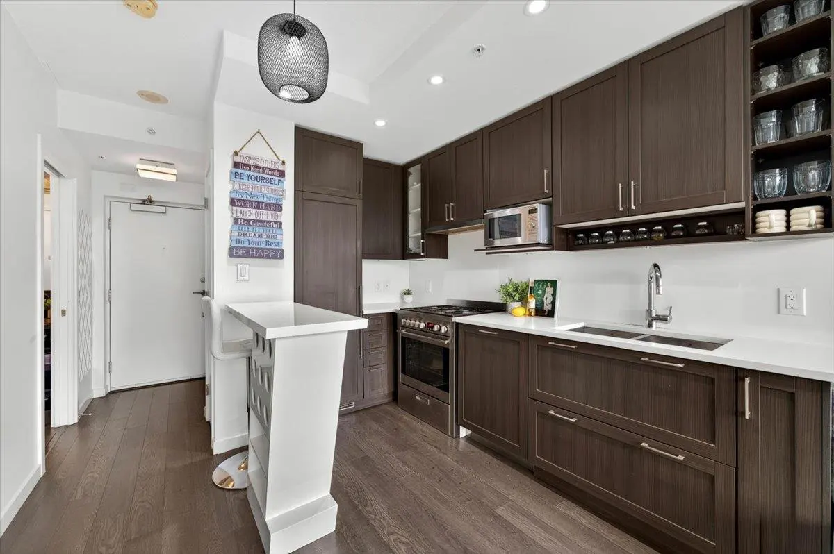 A modern kitchen with dark wood cabinets, white countertops, and stainless steel appliances. A white breakfast bar stands near a colorful wall sign.