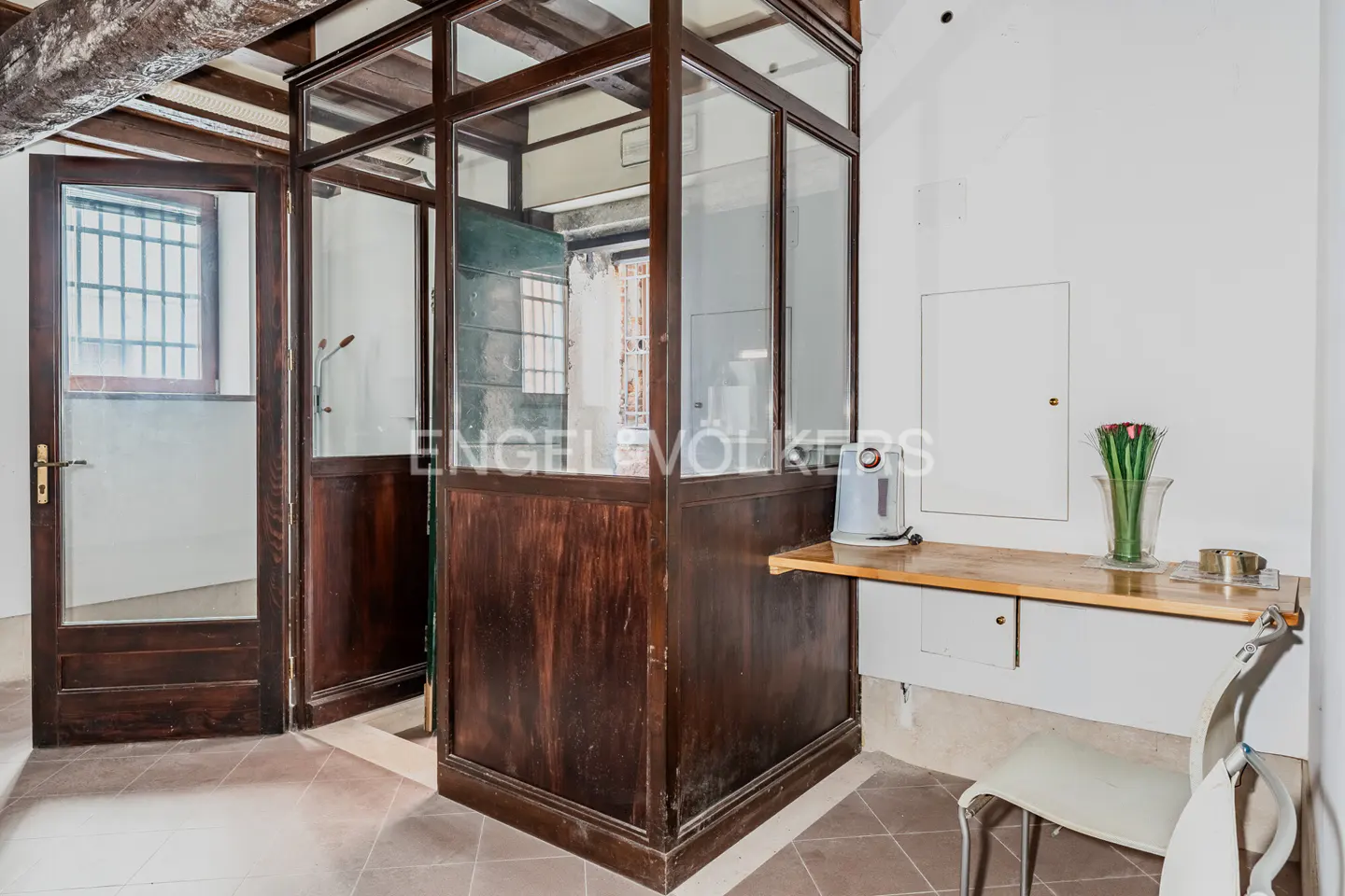 Interior view of a room with a dark wood and glass phone booth, a small table with a kettle, vase, and a chair.