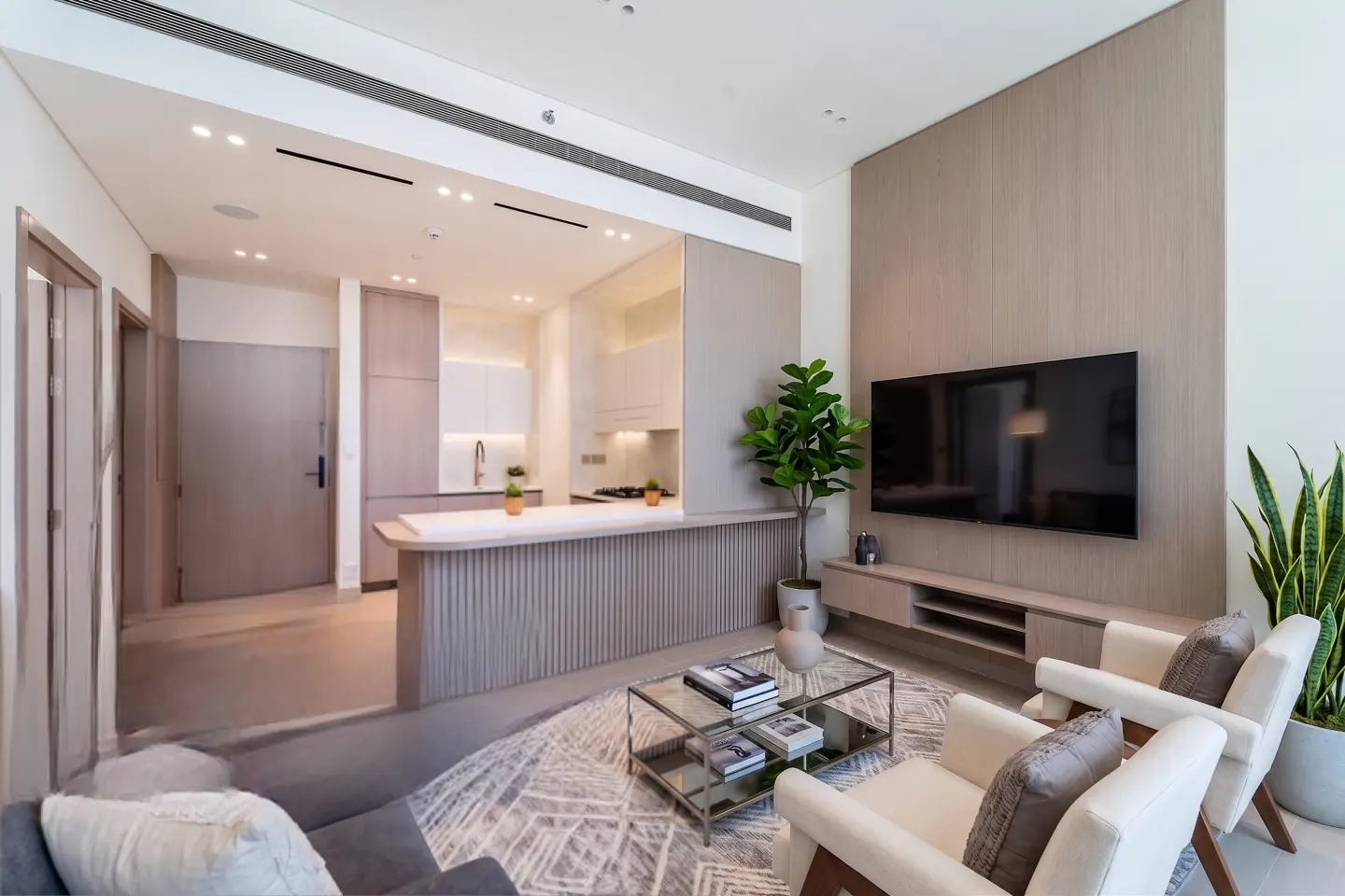 Open-concept living space with a kitchen, two white armchairs, a glass coffee table, and a large TV.