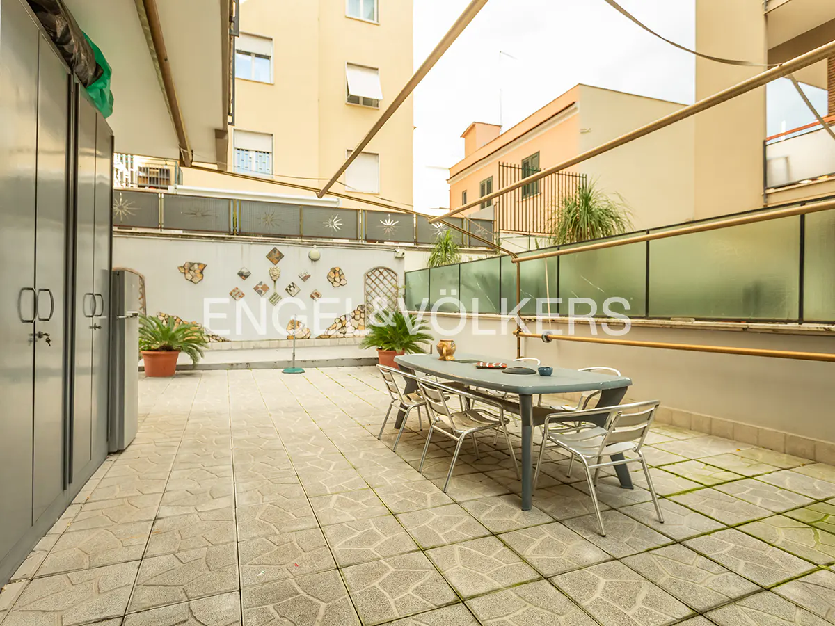 Outdoor patio with a gray table and chairs on a stone tile floor, surrounded by buildings and a decorated wall.