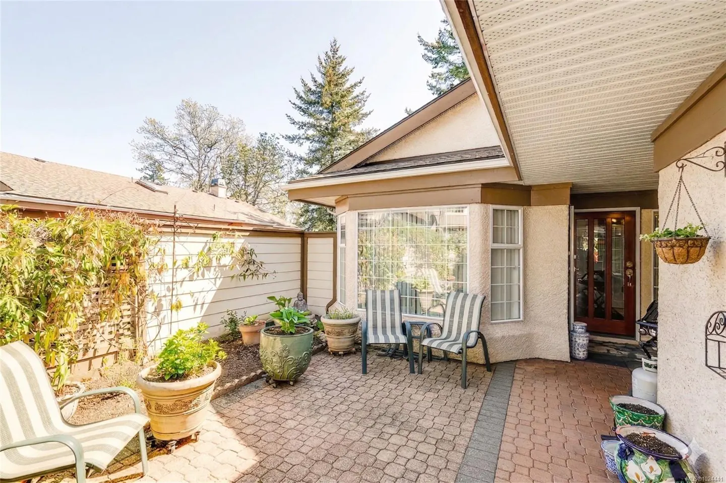 A cozy patio with brick pavers, striped chairs, and potted plants. A bay window and wooden fence add charm.