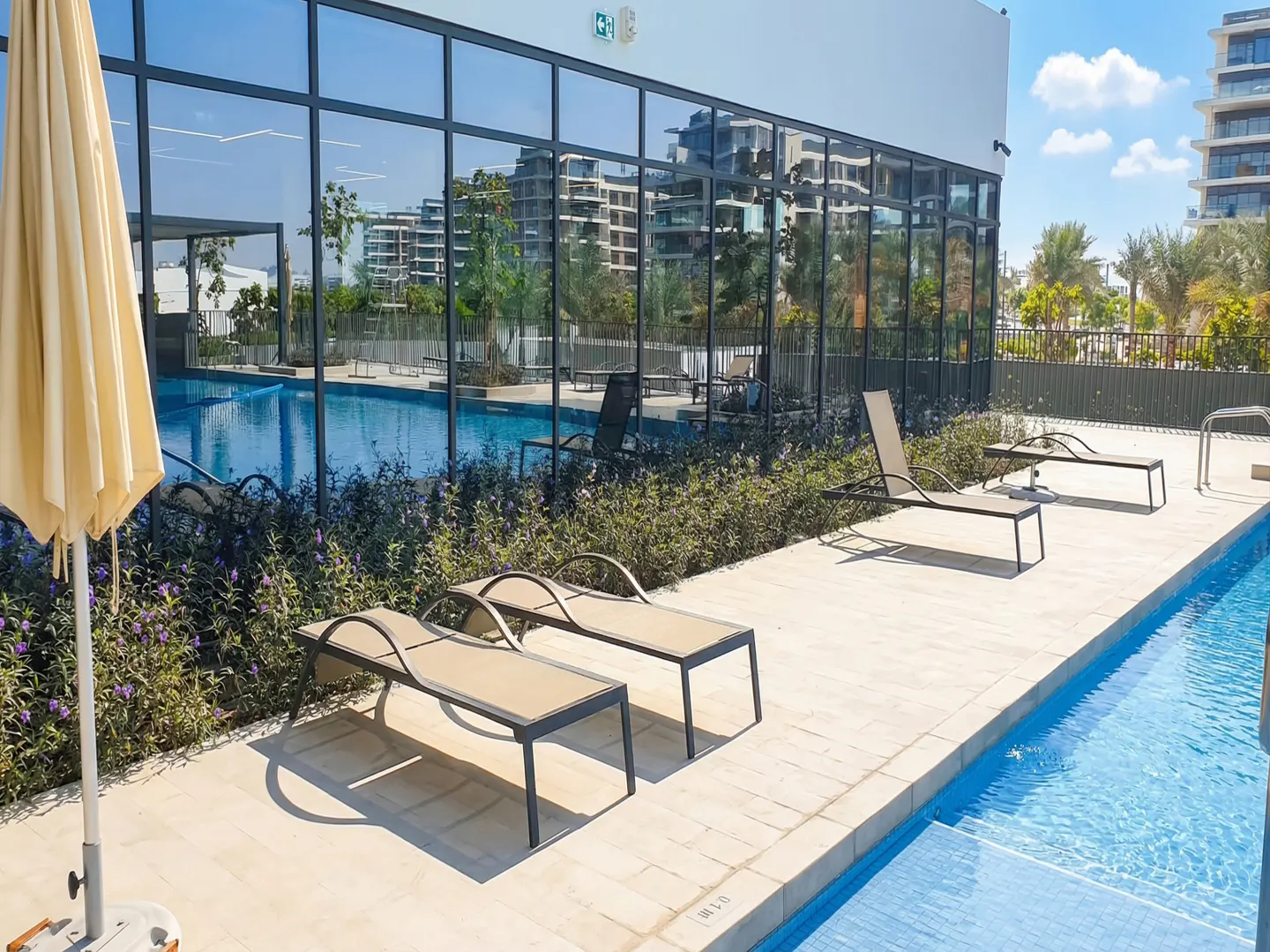 Outdoor pool area with lounge chairs and a beige umbrella. The pool is surrounded by greenery and a building with large glass windows.