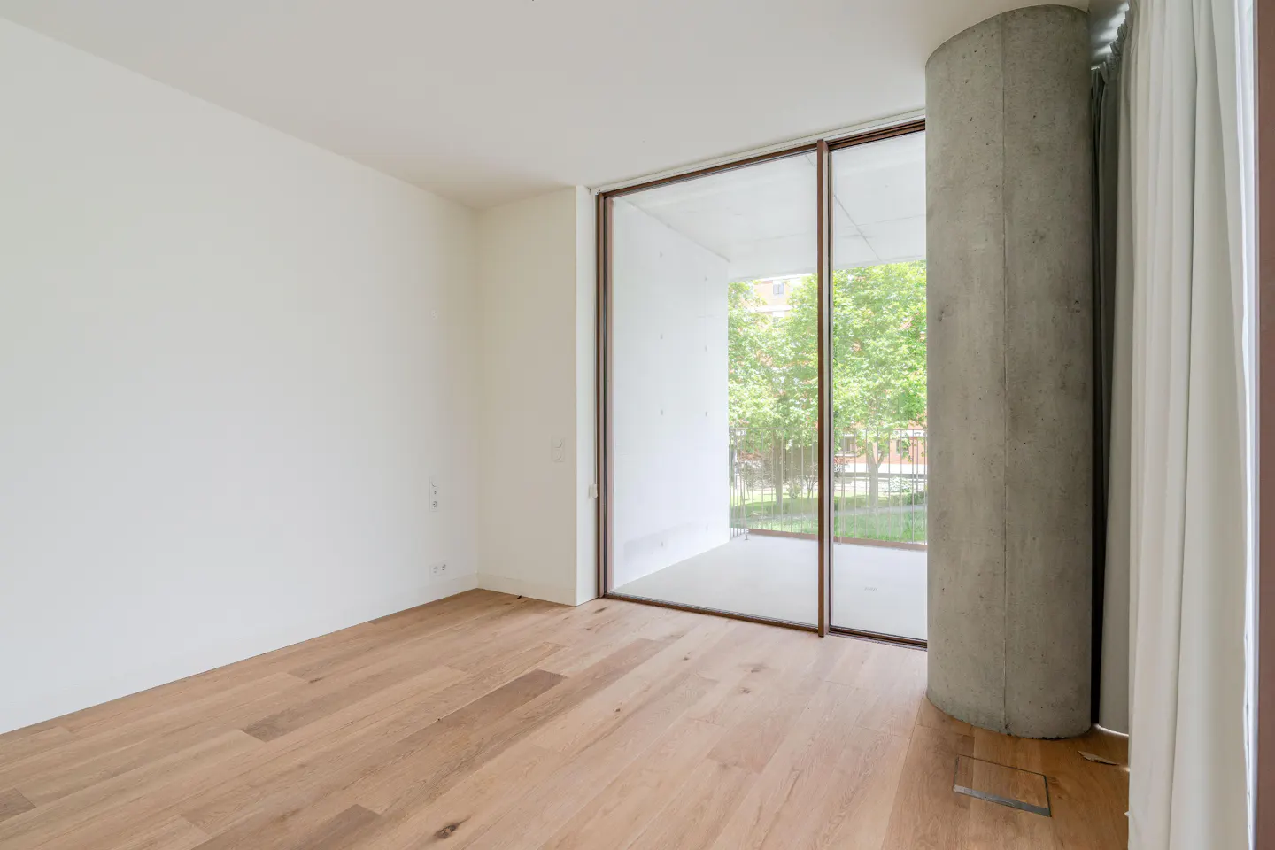 An empty room with light wood floors, white walls, a concrete pillar, and a sliding glass door to a balcony with green trees.