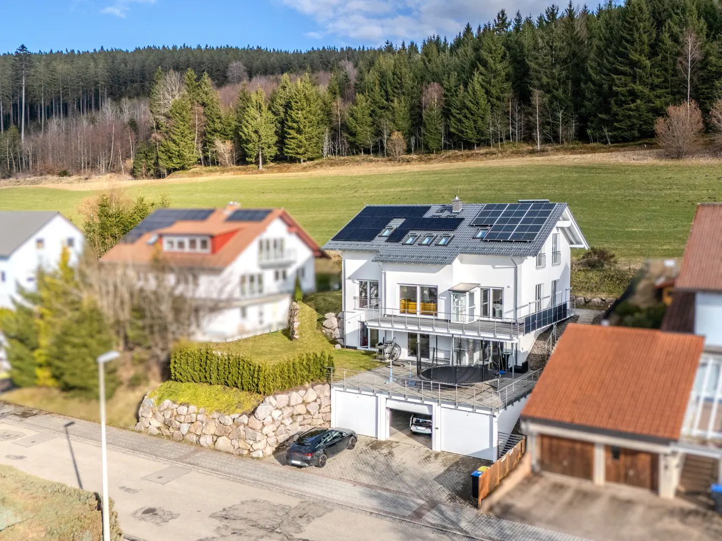 A modern white house with solar panels, a balcony, and a garage, set against a backdrop of green fields and a forest.