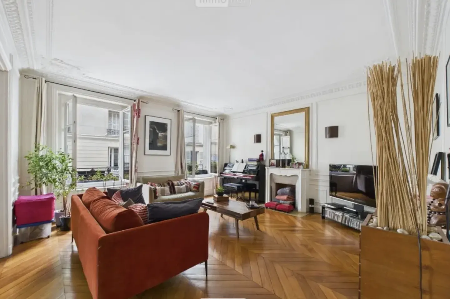 Bright living room with herringbone wood floors, a rust-colored sofa, a piano, fireplace, and large windows.