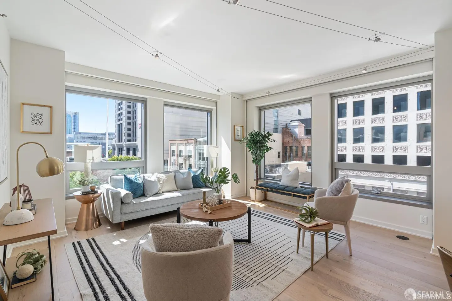 Bright living room with large windows showing city views. A gray sofa, chairs, and a striped rug furnish the space.