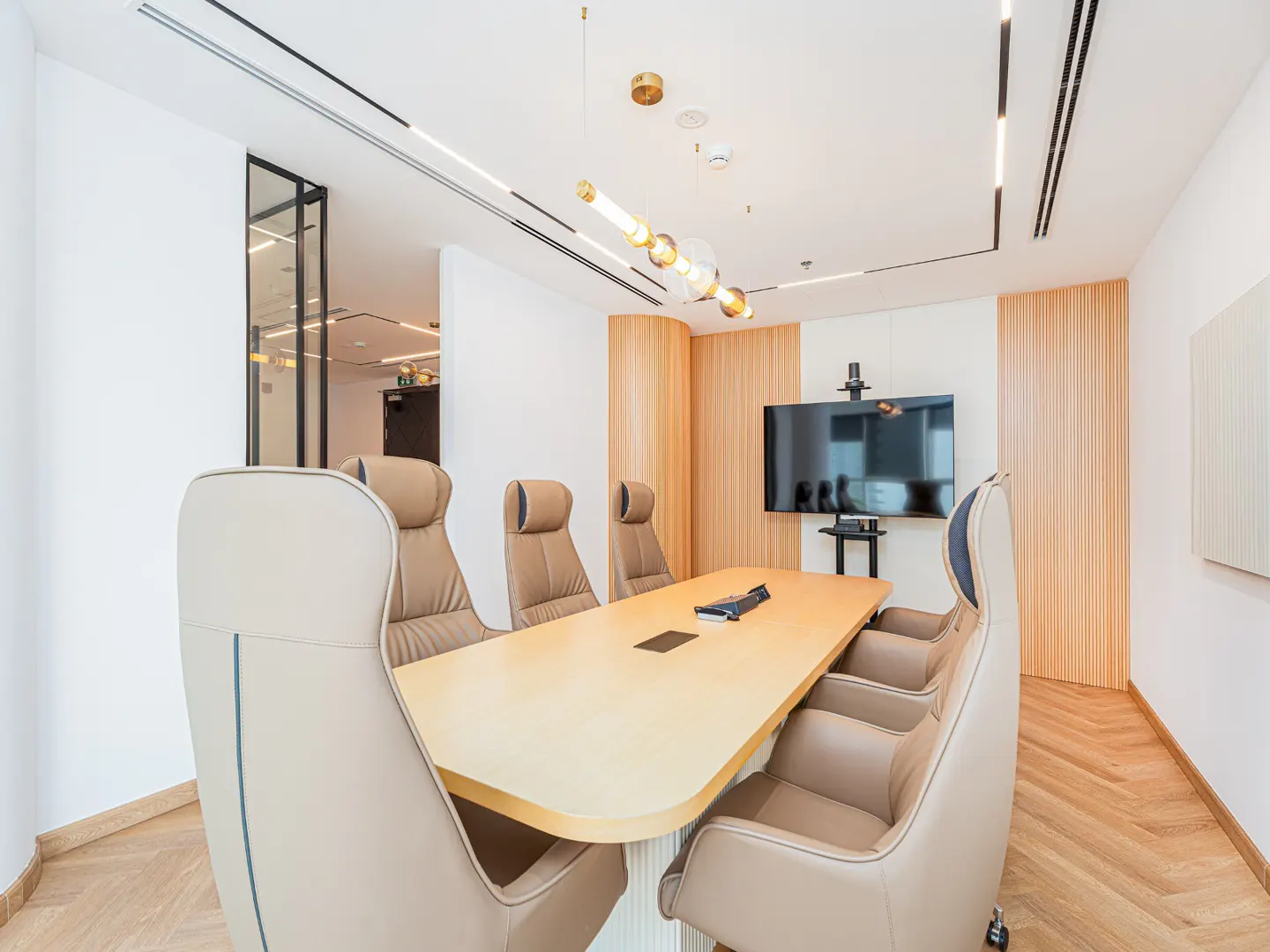 Conference room with a long wooden table, beige leather chairs, and a large TV screen on the wall. Modern lighting fixtures hang from the white ceiling.