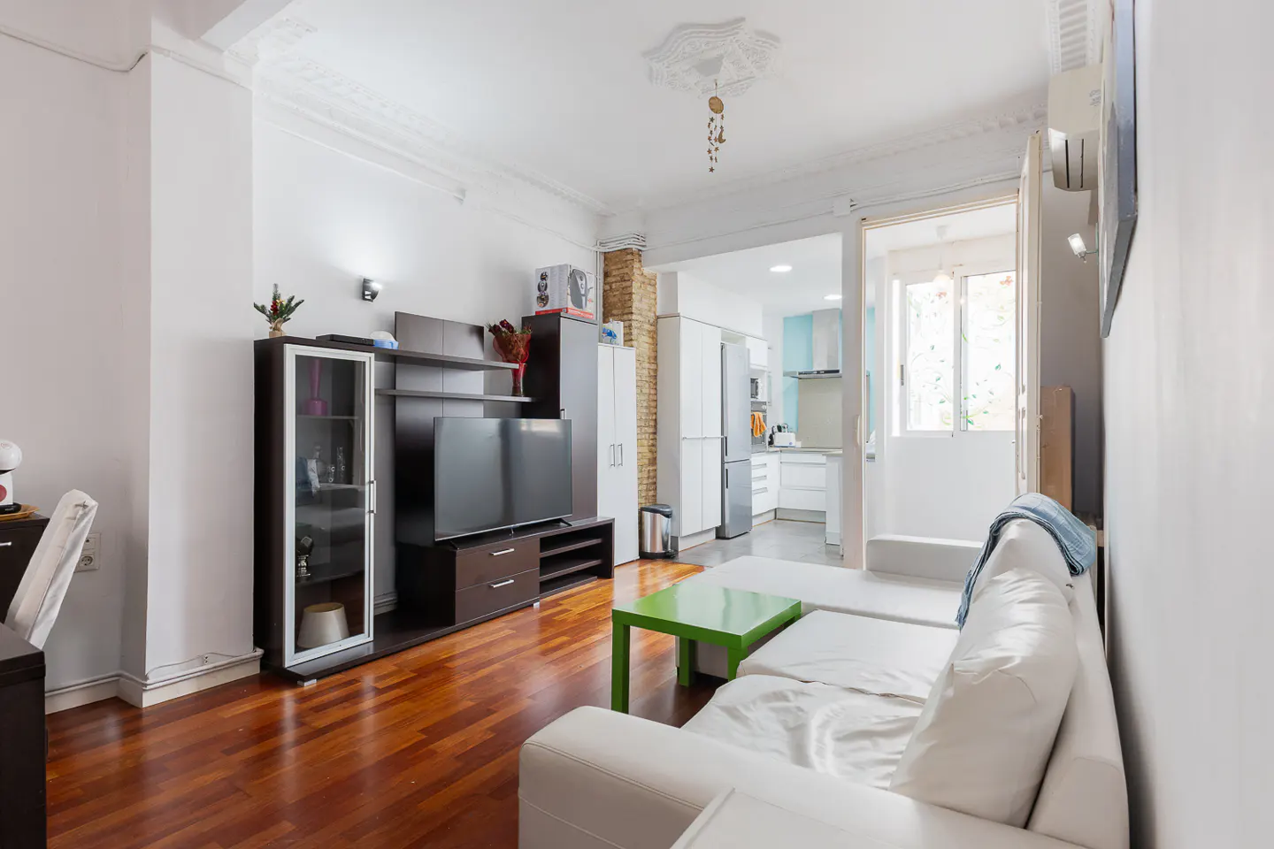 Bright living room with hardwood floors, white walls, and a white sectional sofa. A green table sits in front of a dark entertainment center. Kitchen visible in the background.