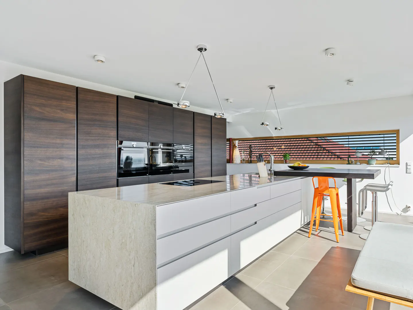 Bright, modern kitchen with dark wood cabinets, white island, and orange stool. Pendant lights hang above the island. A window shows a red tile roof.
