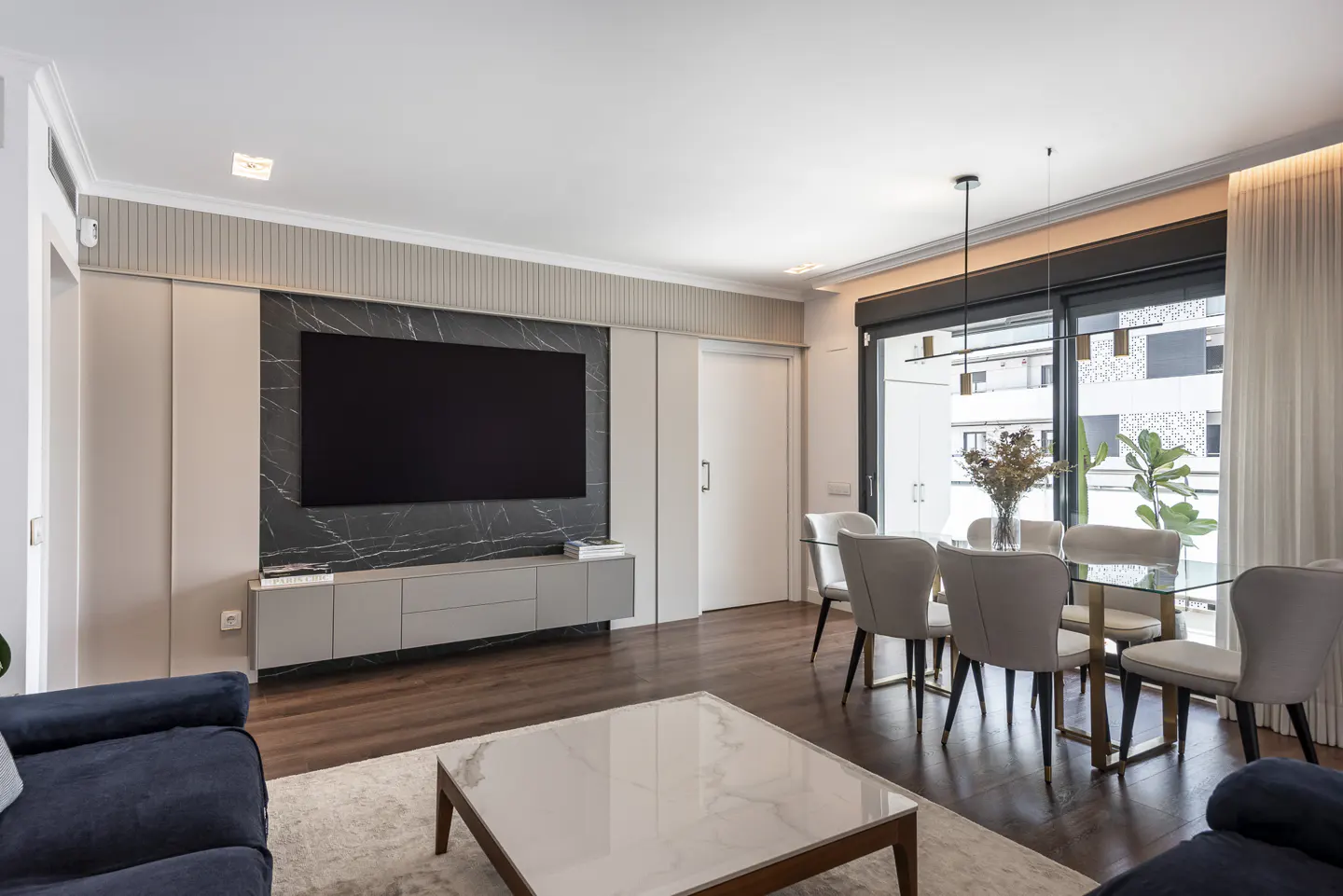 Living room with a large TV on a marble wall, a glass dining table with six chairs, and a coffee table.