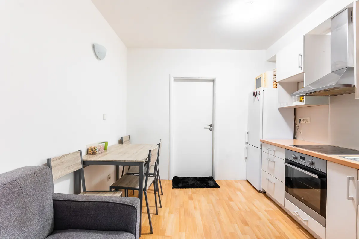 Bright apartment interior with a dining table, gray sofa, and white kitchen cabinets on wood floors.