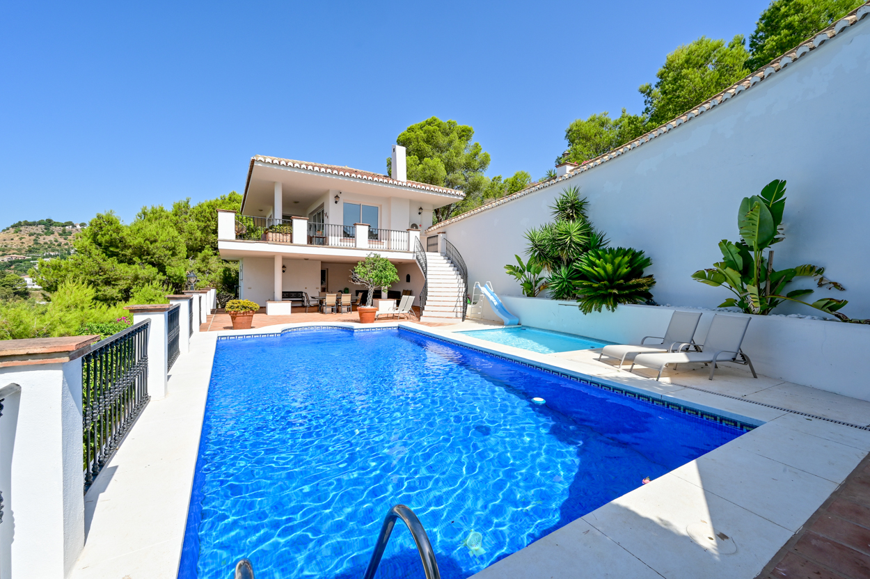 A bright blue swimming pool with a small slide, lounge chairs, and a two-story white villa set against a clear blue sky. [AI-generated text]