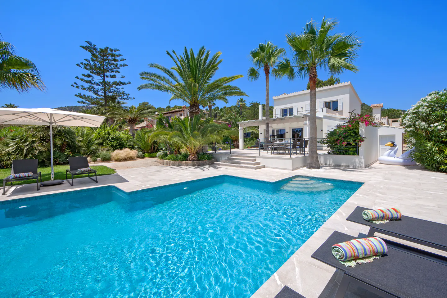 Bright turquoise pool with lounge chairs, palm trees, and a white two-story house under a clear blue sky.