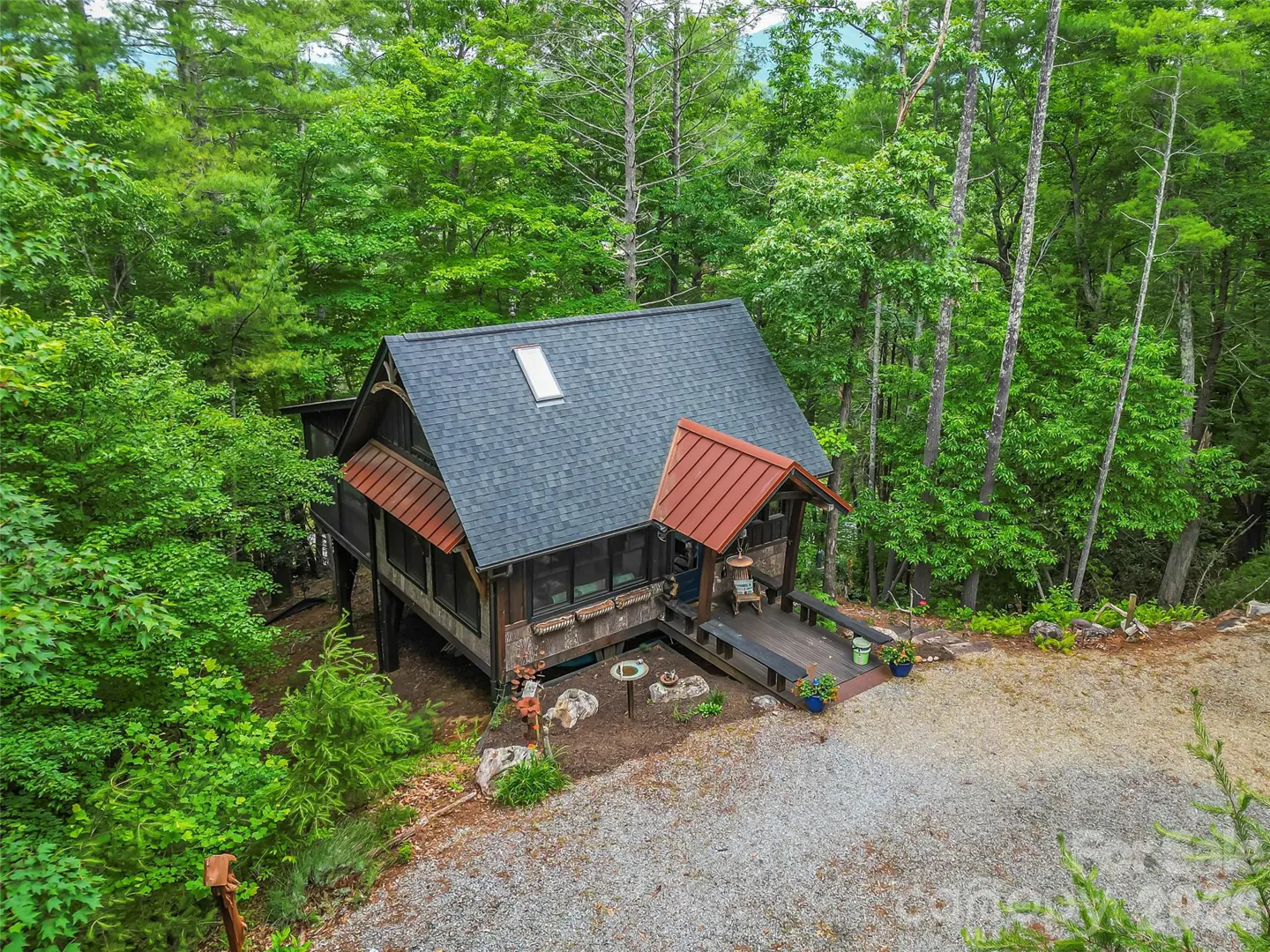 A rustic cabin with a dark shingled roof and red metal accents, nestled in a lush green forest with a gravel driveway.