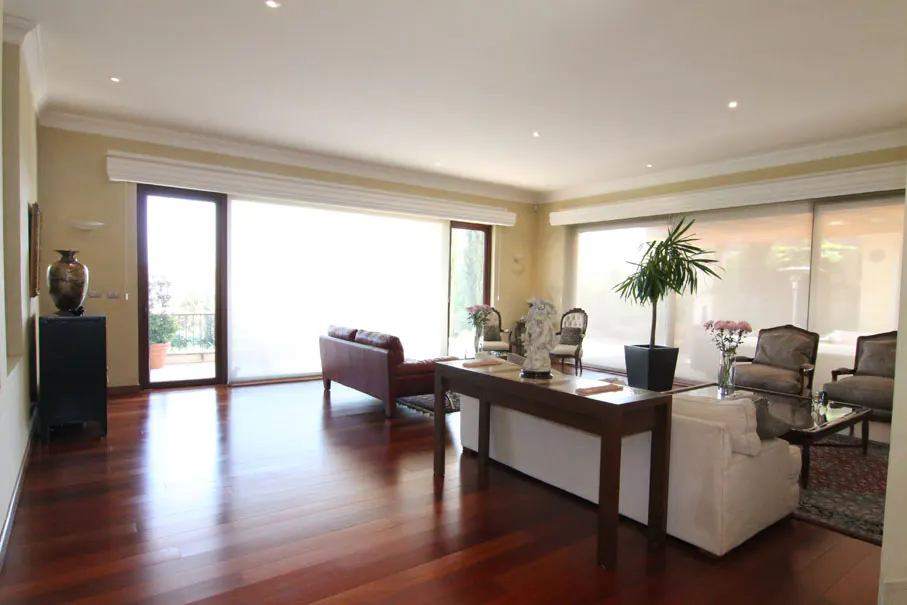 Bright living room with hardwood floors, large windows, and neutral-toned furniture. A brown leather sofa and a white sofa are visible.