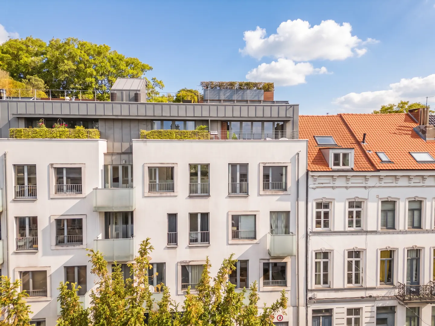 Exterior view of white apartment buildings with balconies and rooftop gardens under a blue sky with white clouds.