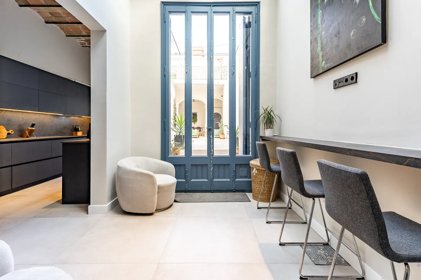 Interior view of a modern kitchen and dining area with blue doors leading to a courtyard. Gray chairs line a counter, and a cream-colored chair sits nearby.