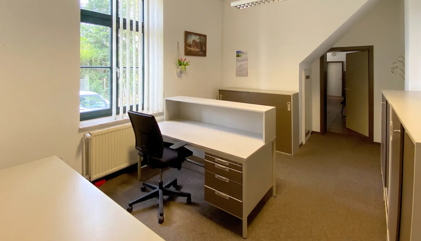 Office space with a white desk, black chair, and brown cabinets. A window with blinds is to the left, and a doorway leads to another room.