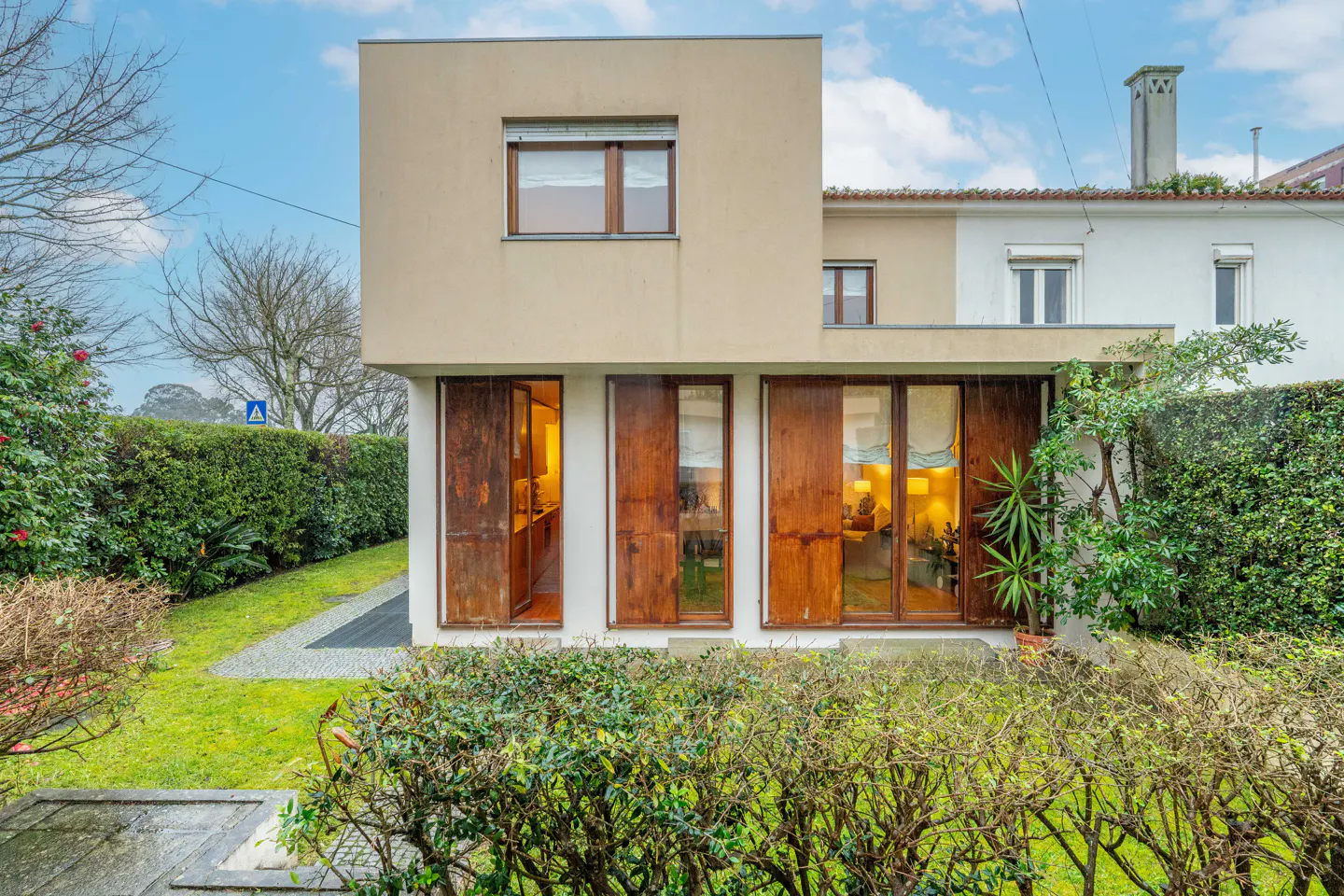 Modern two-story house with large wooden doors and green lawn. The house is beige with a square design and a small window on the second floor.