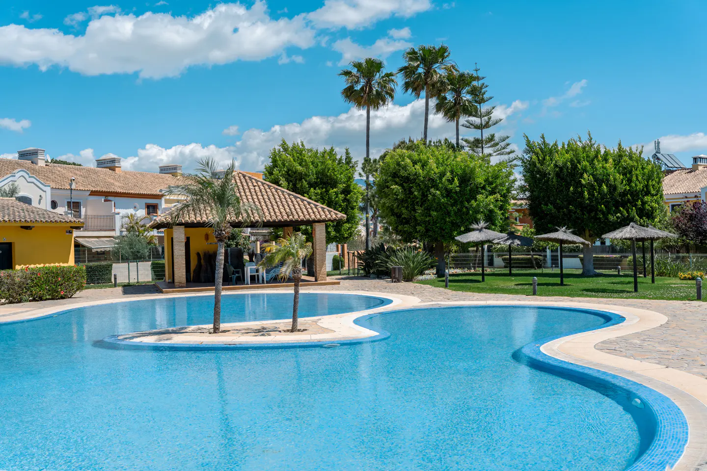 A bright blue swimming pool with a small island, surrounded by palm trees, green lawns, and buildings under a blue sky.