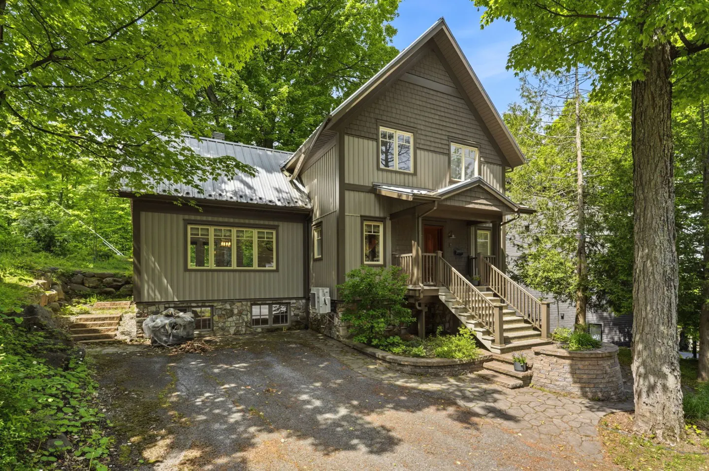 Two-story taupe house with a metal roof, stone foundation, and wooden porch, surrounded by lush green trees.