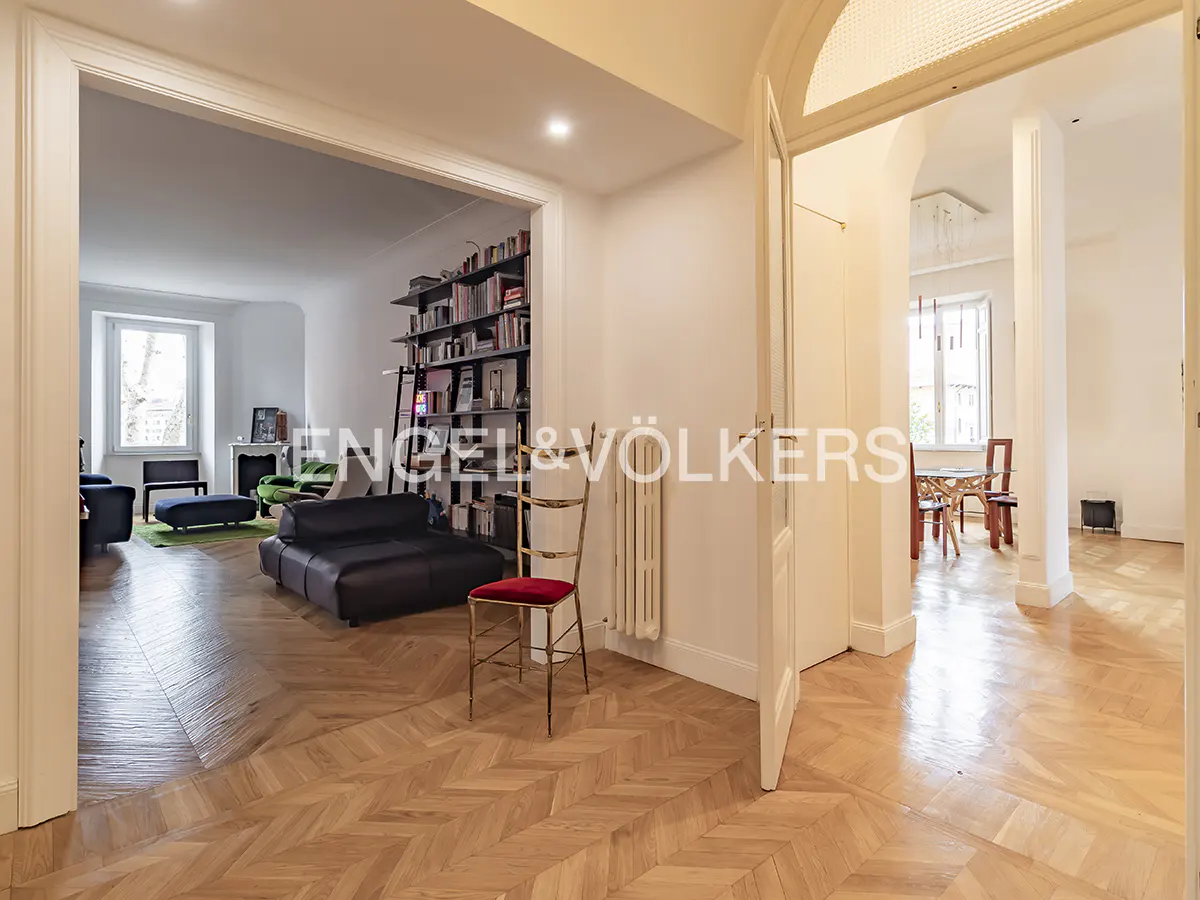 Open doorway reveals a living room with a black sofa, bookshelf, and herringbone wood floors, plus a dining room beyond.