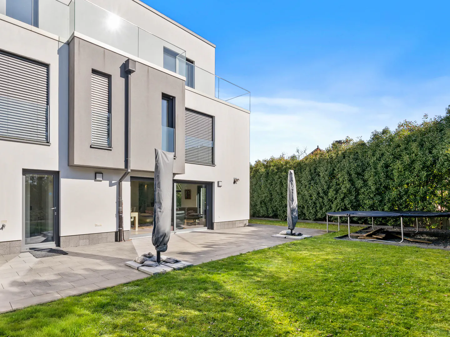 Modern two-story home with a patio, green lawn, and trampoline. The house is white with gray accents and has large windows.