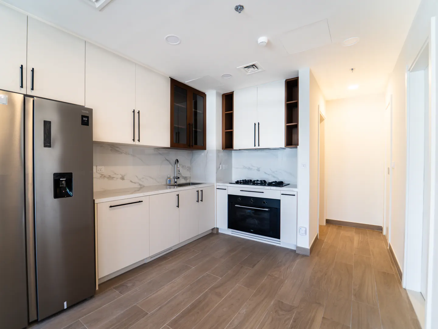 A modern kitchen with white cabinets, black hardware, stainless steel fridge, and wood-look tile flooring.
