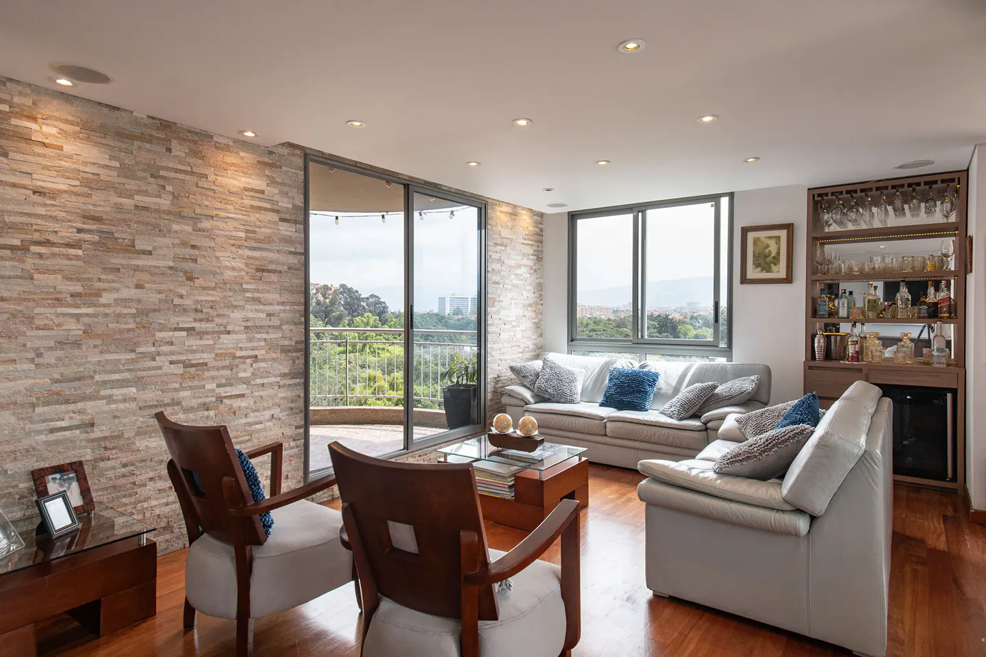 Living room with stone wall, wood floors, and gray sofas. Balcony with city view. Bar with glasses and bottles.