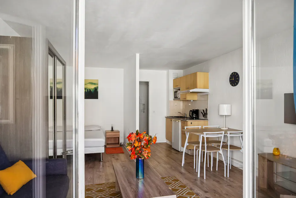A bright studio apartment with a kitchenette, dining table, and bed. A vase of orange flowers sits on a coffee table.