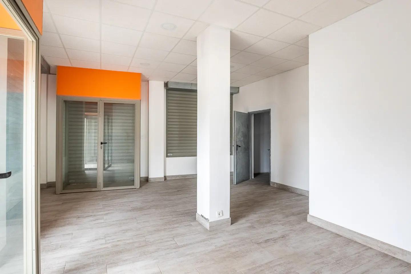 Empty commercial space with white walls, gray tile floor, and a large orange sign above the glass storefront. A white pillar stands in the center of the room.