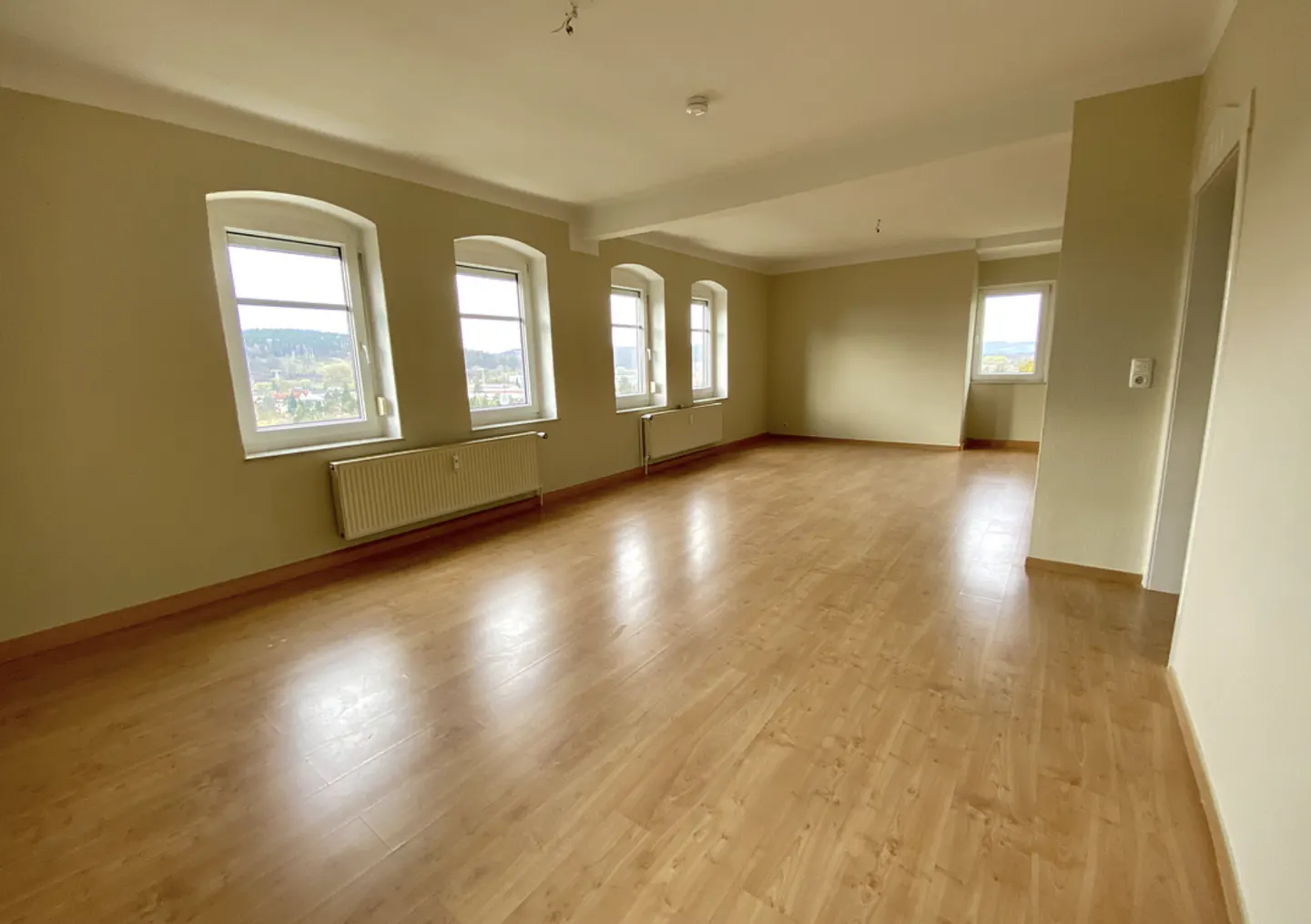 Empty room with wood floors, light beige walls, and white trim. Three arched windows with white frames overlook a town. Radiators under windows.