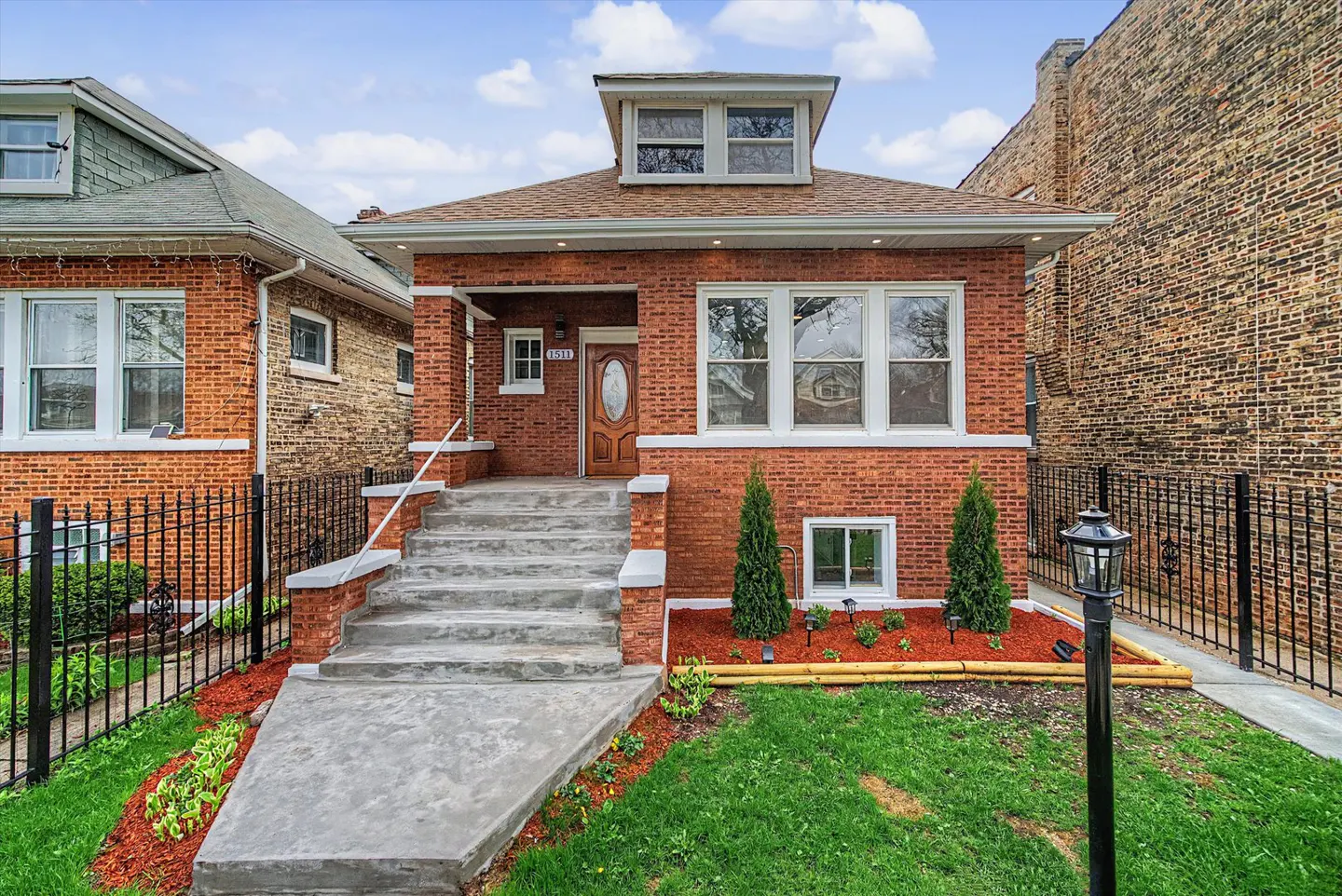 Red brick house with concrete steps leading to a wood door. A black metal fence surrounds the green lawn.