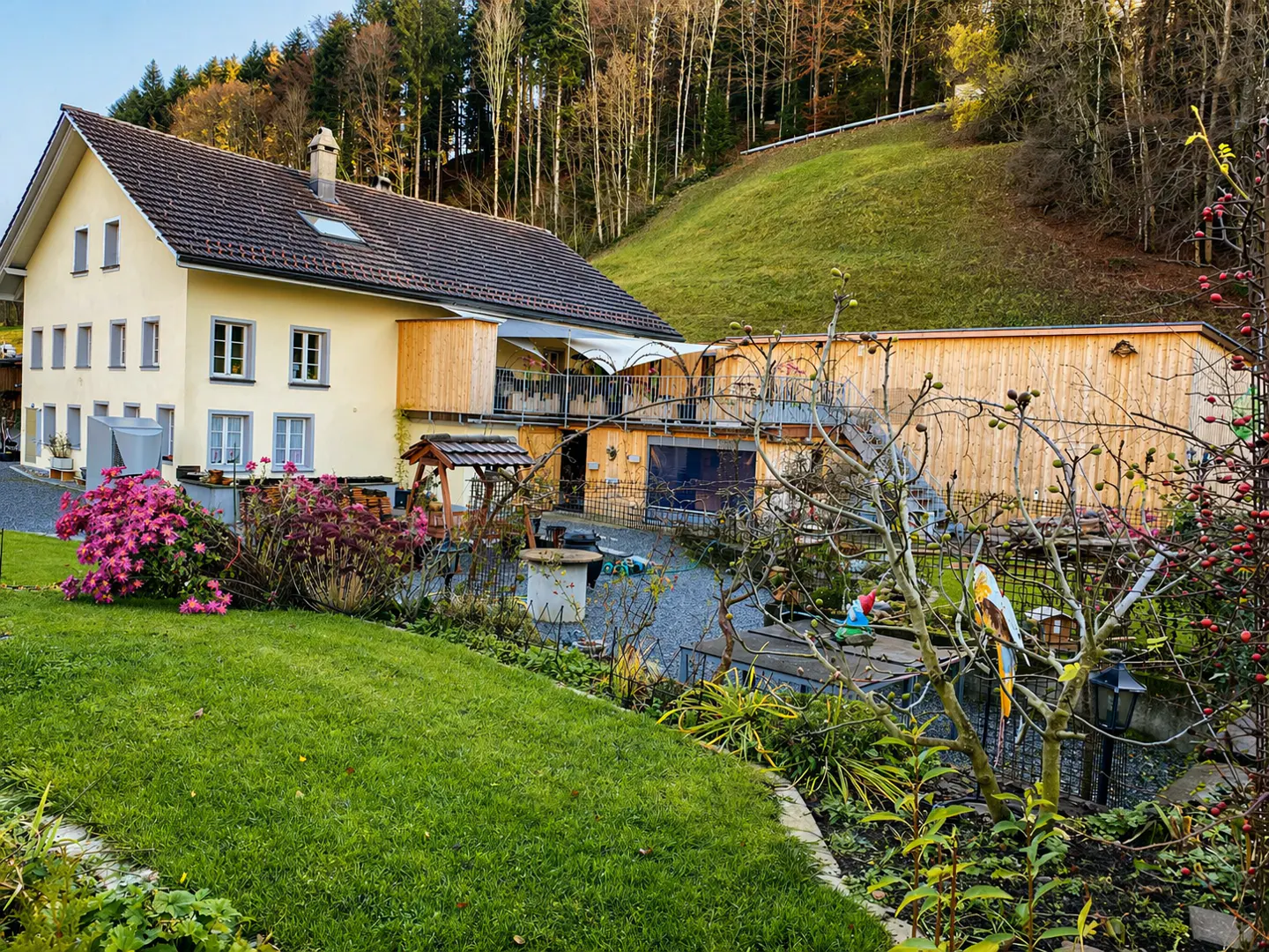 Exterior view of a yellow two-story house with a brown roof, a green lawn, and a wooded hillside in the background.