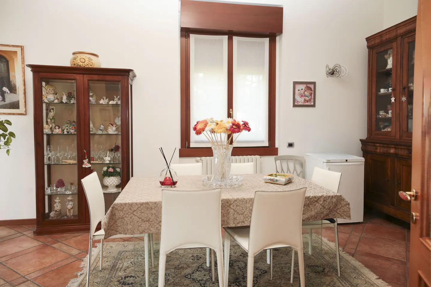 A dining room with a table, white chairs, a brown cabinet, and a window with white curtains.