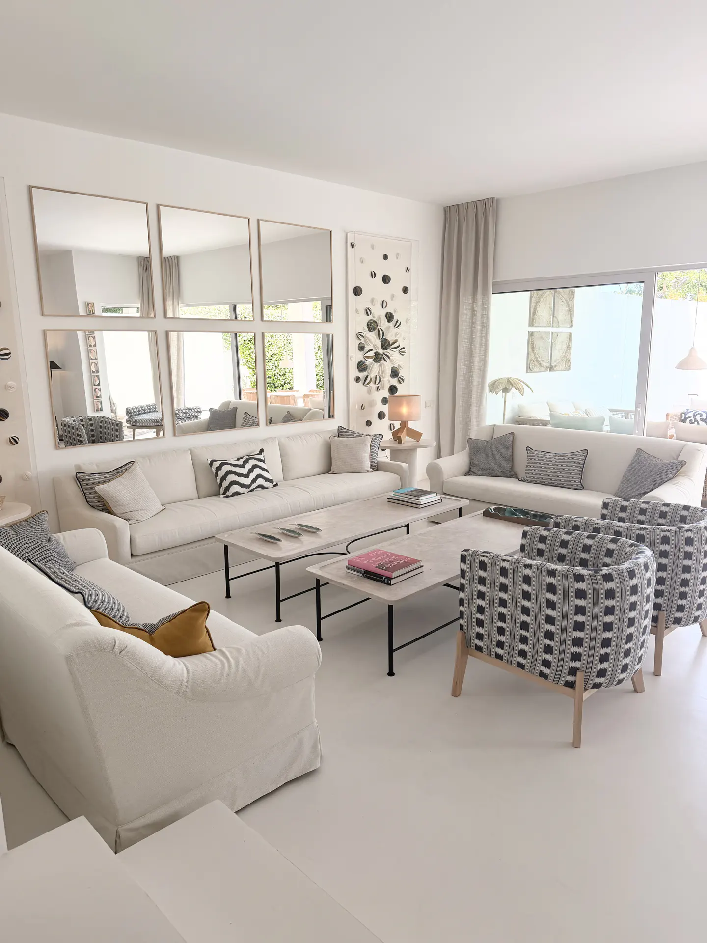 Bright living room with white sofas, patterned chairs, and square mirrors on the wall. Coffee tables and a large window add to the airy feel.