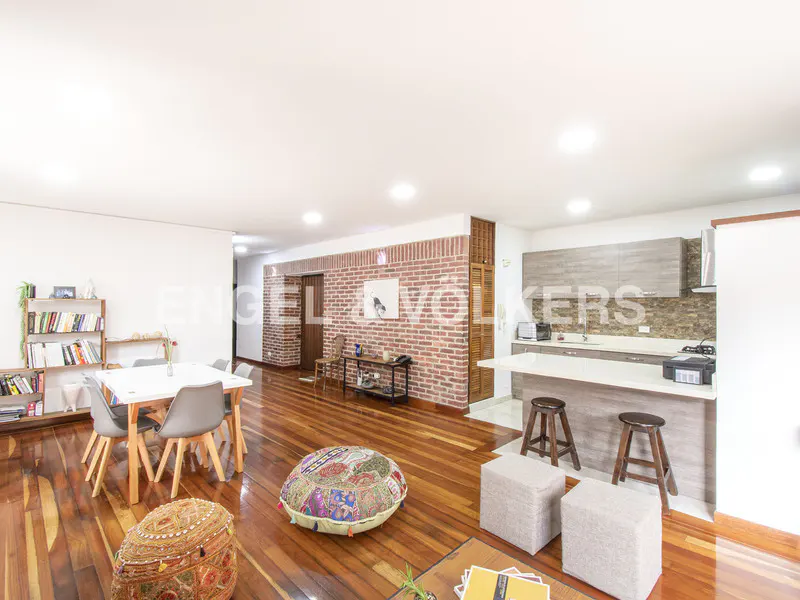 Open-concept living space with hardwood floors, a dining table, and a kitchen island with bar stools. A brick wall adds texture.
