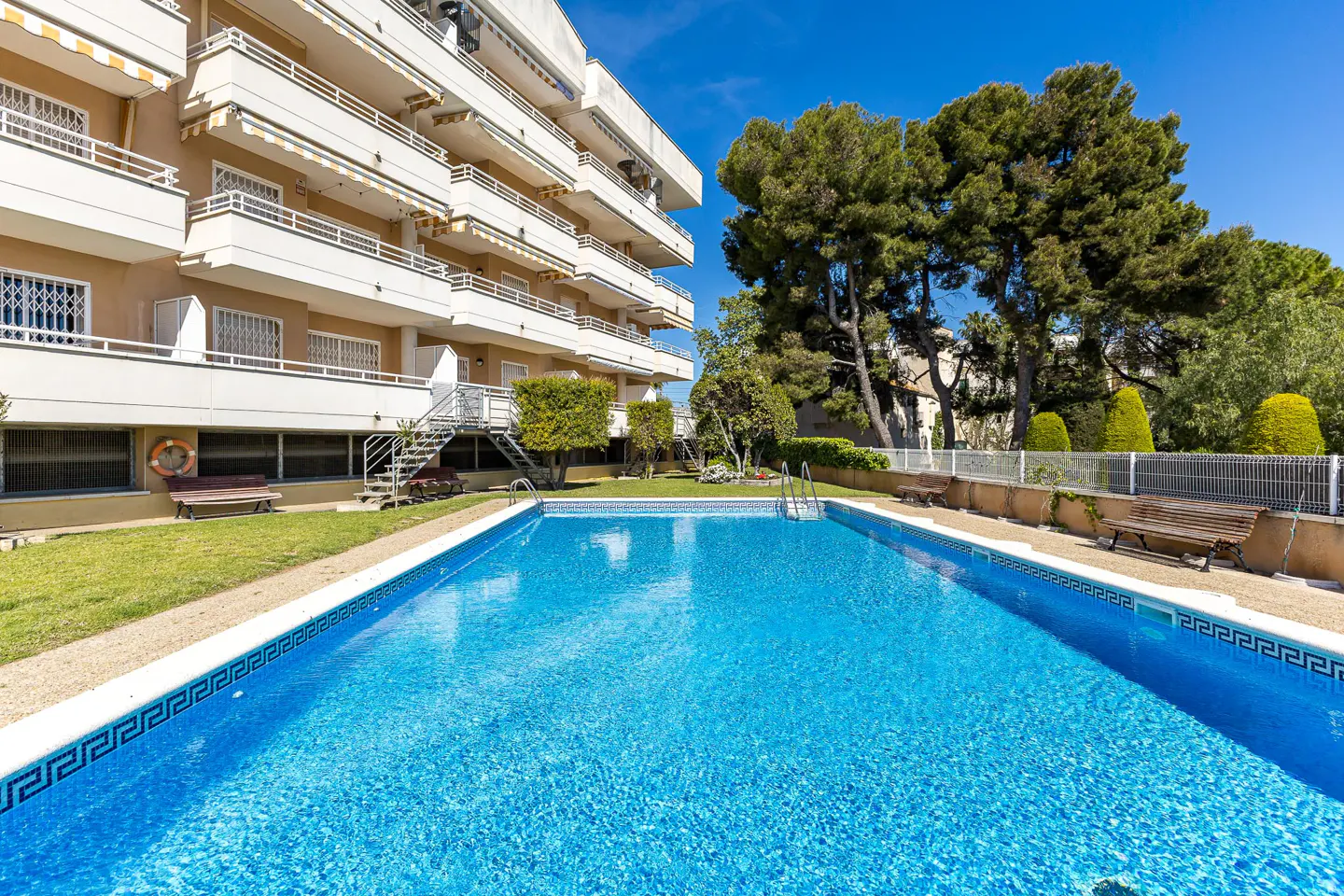 Exterior view of a blue tiled swimming pool in front of a multi-story apartment building with balconies and green trees.