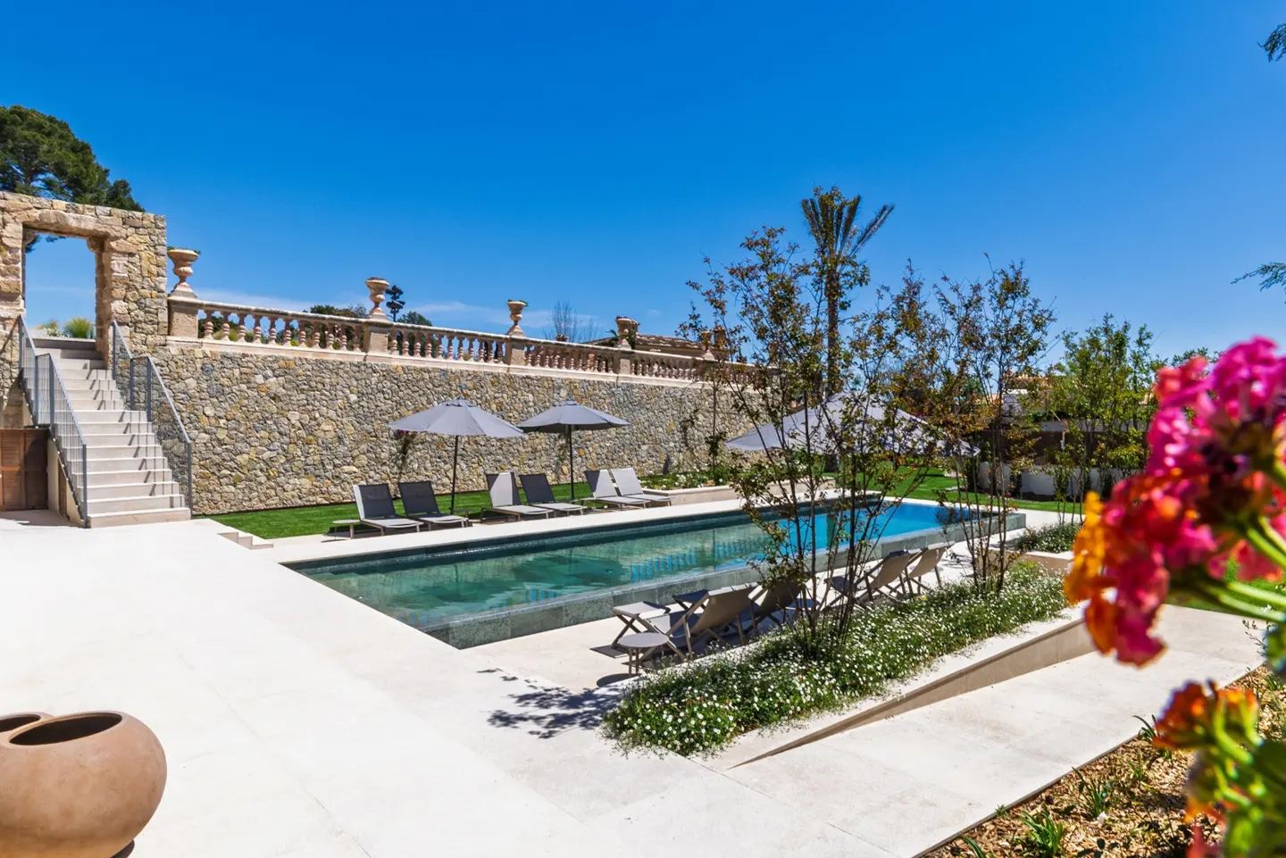 Luxury pool area with lounge chairs, umbrellas, and stone wall. Bright blue sky and colorful flowers in the foreground.