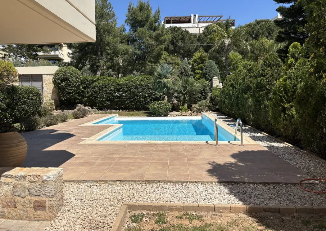 A backyard swimming pool with blue water, surrounded by brown tiles and white gravel, with green trees in the background.
