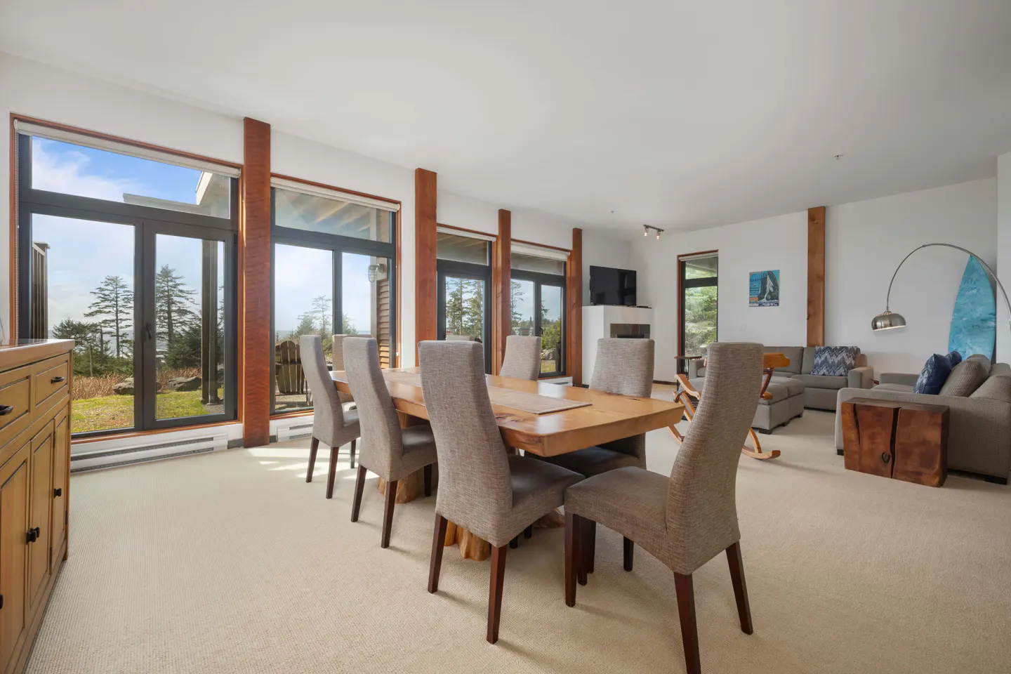 Bright, open-concept living space with a wood dining table, upholstered chairs, and large windows overlooking a green landscape.