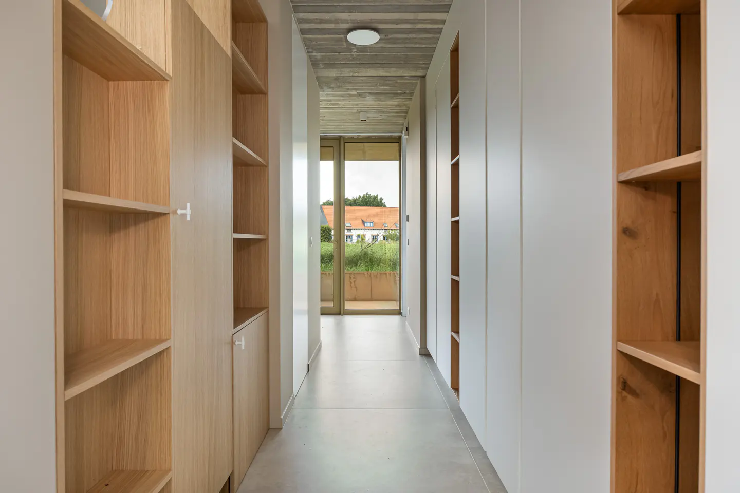 Modern hallway with light wood shelving, white cabinets, a textured concrete ceiling, and a view of a garden through glass.