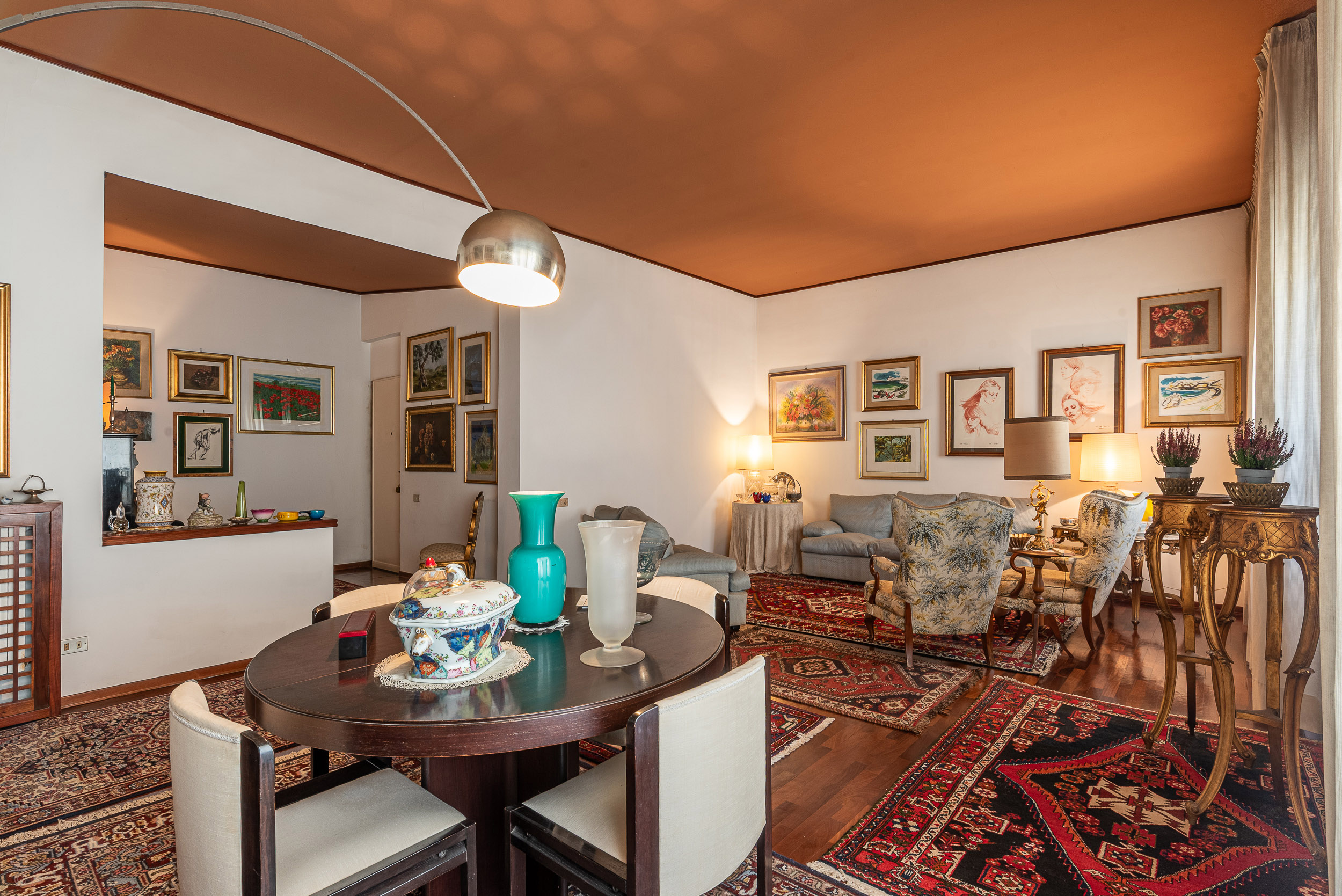 Living room with a dark wood dining table, patterned rugs, traditional armchairs, and walls filled with framed artwork. [AI-generated text]