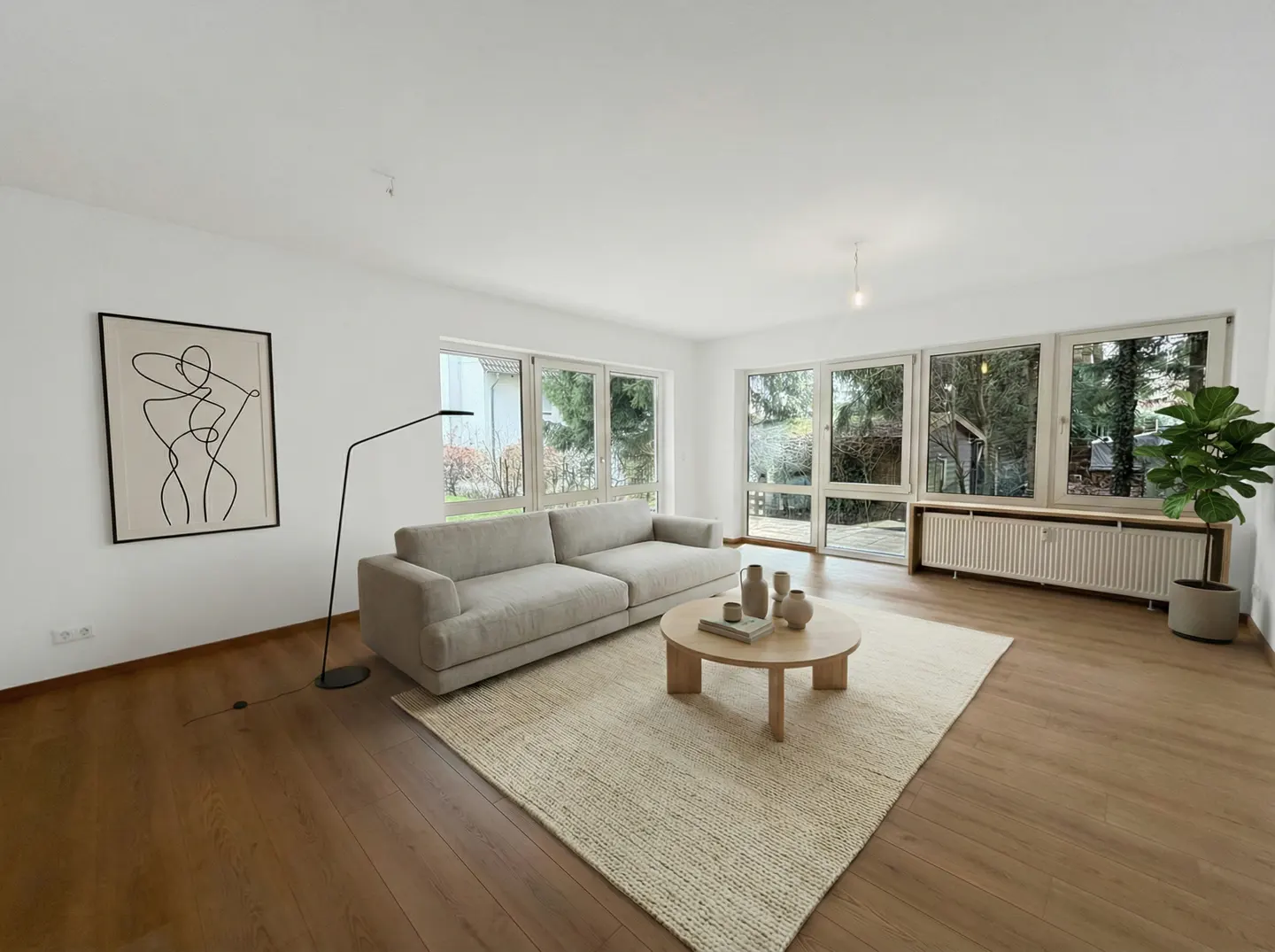 Bright living room with wood floors, white walls, and large windows. A beige sofa sits on a woven rug, with a round coffee table. Modern art and a floor lamp accent the space.