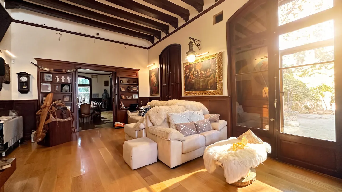 A sunlit living room with wood floors, beams, and trim. A cream sofa with pillows and a fur throw sits near large doors to a garden.