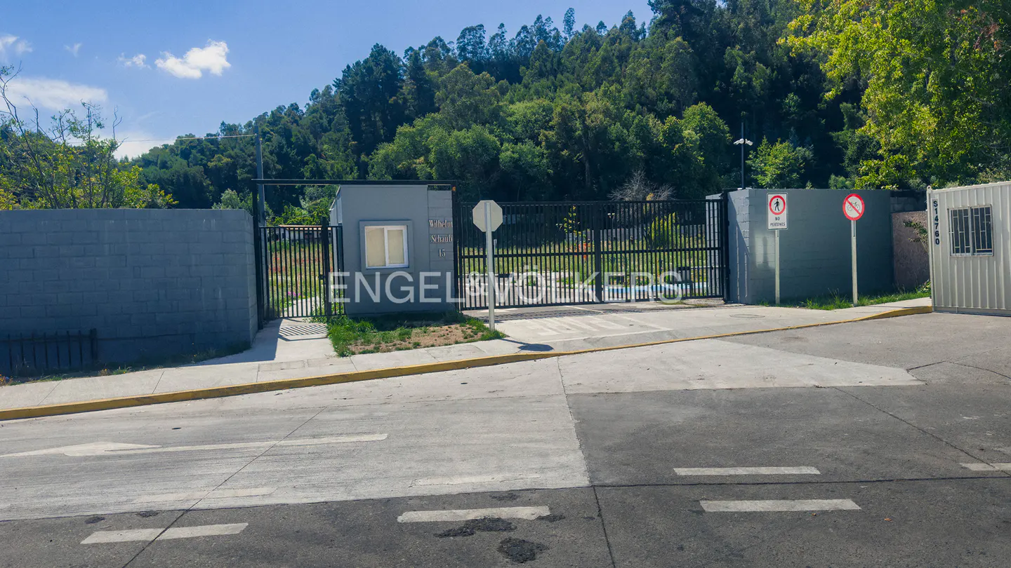Entrance to Engel & Volkers property with black metal gate, gray walls, and guard house. Trees in background.