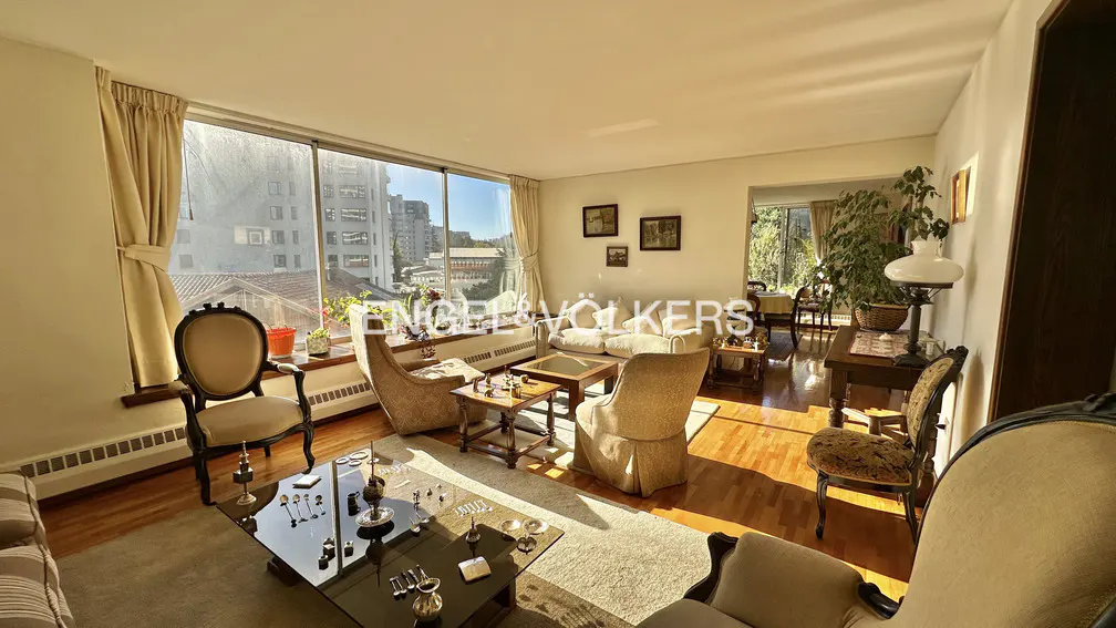 Sunlit living room with hardwood floors, beige furniture, and a large window overlooking a city view.