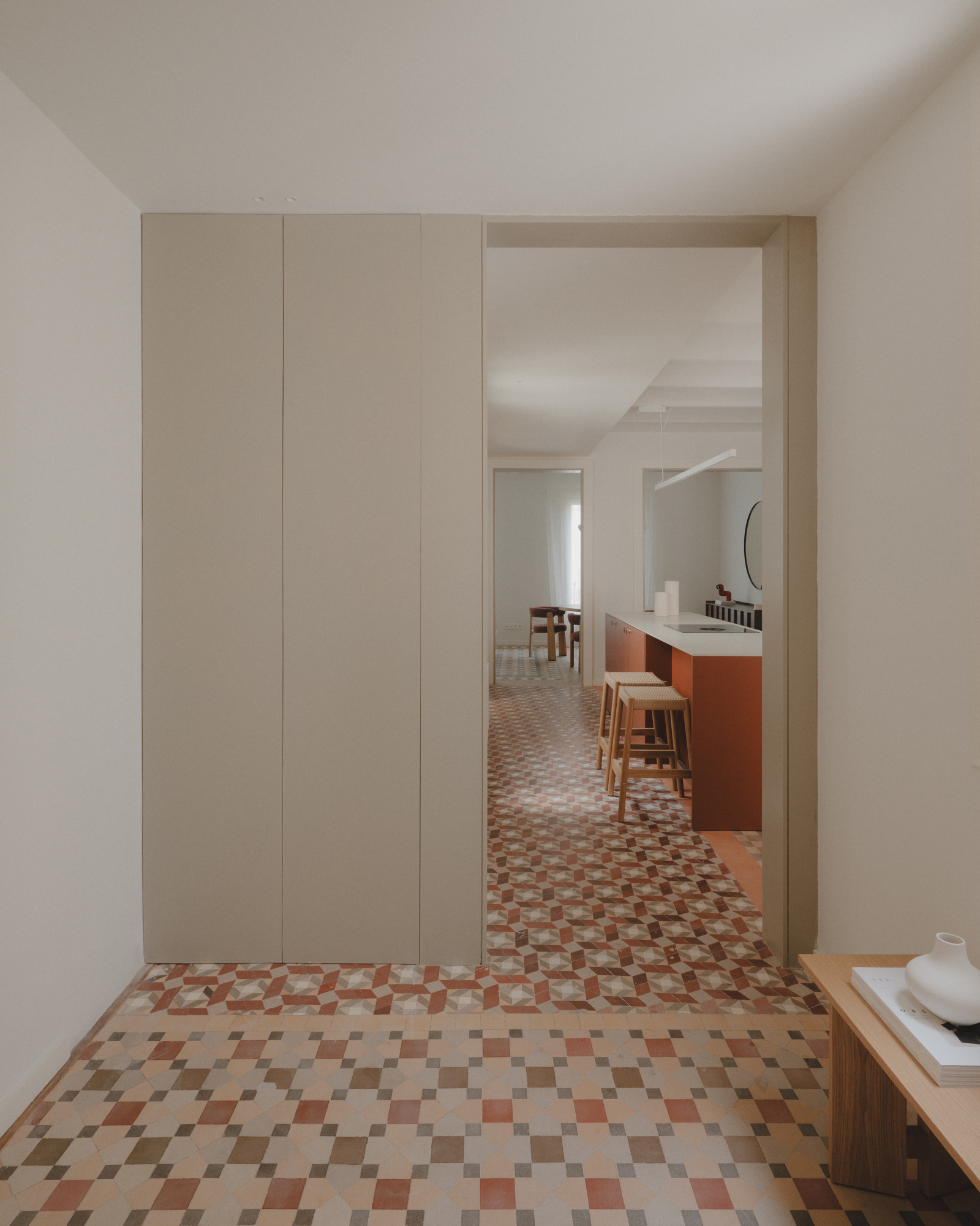 Minimalist room with beige wall panels, patterned tile floors, and a view into a kitchen with a terracotta island.